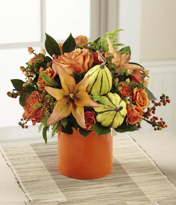 Swirling orange roses, orange spray roses, and star-shaped peach Asiatic Lilies are surrounded with the eye-catching textures of yellow solidago, bittersweet stems, aralia leaves and lush greens with brilliant yellow gourd accents tucked in a just the right spot, all beautifully arranged in an orange ceramic cylinder vase. STANDARD SIZE