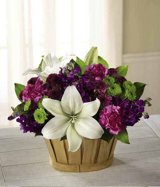 White Asiatic Lilies are clean and bright against a berry colored back drop of purple gilly flower, hot pink carnations, green button poms, purple button poms, lavender roses, and lush greens. Arranged lovingly in a rectangular woodchip baske