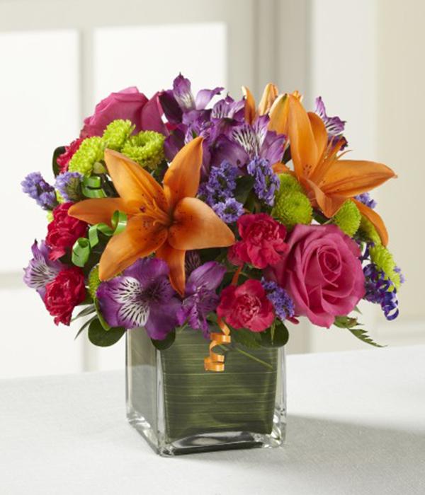 Hot pink roses and orange Asiatic Lilies are vibrant and fun surrounded by purple Peruvian Lilies, hot pink mini carnations, green button poms, purple statice, and an assortment of lush greens. Accented with assorted curling ribbons to give it that party feel and presented in a clear glass cubed vase lined with ti leaf green material 