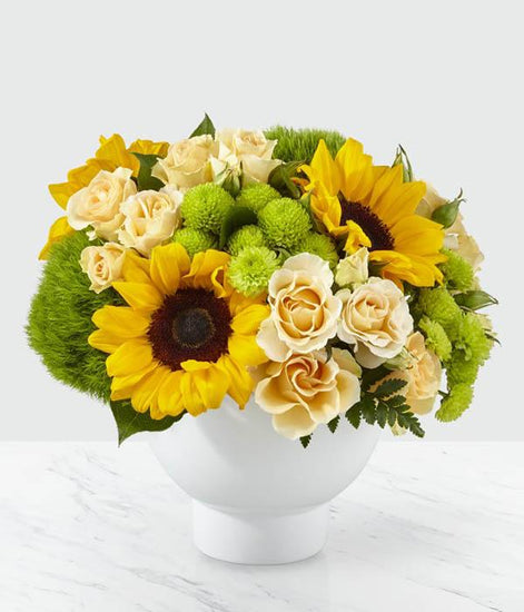 From the bold beauty of bright sunflowers to the sweet delight of peach spray roses. Complete with bursts of green trick dianthus and button pompons, this arrangement is set in a modern white ceramic vase