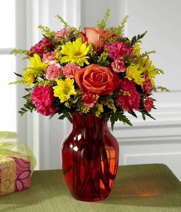 Orange roses, yellow daisies, hot pink carnations, orange mini carnations, yellow solidago, and lush greens are arranged beautifully in an eye-catching orange glass vase.