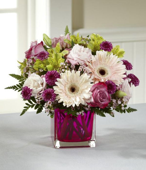  pale pink gerbera daisies, lavender roses, pale pink double lisianthus, green Peruvian Lilies, clusters of pink waxflower, and burgundy button poms. Accented with lush greens and arranged perfectly in a modern raspberry glass cubed vase