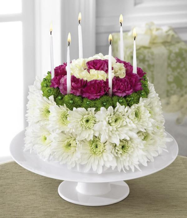 Perfectly arranged in the shape and styling of a colorful birthday cake are white chrysanthemums, green button poms, pale yellow carnations and magenta mini carnations. Presented on a white cake plate, this memorable flower arrangement will add to the festivities of their special day.