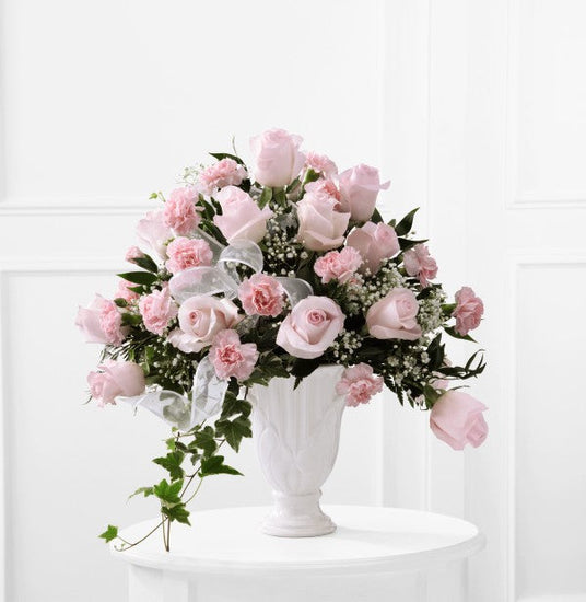 Pale pink roses and pink carnations are offset by baby's breath, variegated ivy, lush greens, and a white sheer ribbon, gorgeously arranged in a designer white ceramic pedestal vase