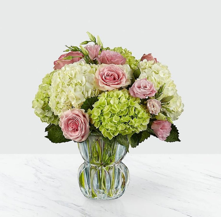  The blushing swirl of the roses and lisianthus bring a sweet drama to this arrangement offset by clusters of green and cream hydrangea. With the vase tinted in a subtle blue shade